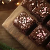 Fotografia vista de cima de vários quadrados de brownie com confeitos de natal, sobre uma tábua de madeira que está apoiada em uma mesa de madeira que tem luzes de natal.