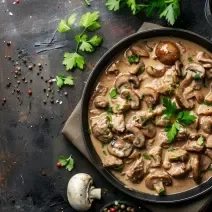 Fotografia vista de cima de uma frigideira de ferro com o molho madeira com champignon. Por cima tem algumas folhas de salsinha. A frigideira está sobre um pano marrom que está apoiado em uma mesa de madeira, com grãos de pimenta e folhas de salsinha