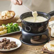 Fotografia em tons de marrom e beje de uma bancada de madeira com uma panela de fondue preta com fondue de queijo. Ao lado um prato redondo com carne, um prato retangular com batatas e brócolis, e uma cesta com pães.