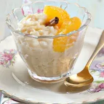 fotografia em tons de branco e amarelo tirada de um prato redondo branco com um copo transparente que contém arroz doce com rodelas de laranja de canela