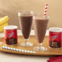 Foto em tons de vermelho da receita de shake saudável feita com Nescau Extra Cacau servida em dois copos altos de vidro sobre uma tábua de madeira com duas latas de nescau e canudos listrados. Ao fundo, bananas e morangos para decorar  sobre uma mesa bege