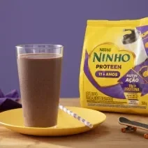 Fotografia de um copo de vidro grande com um smoothie de açaí apoiado em um prato amarelo raso com um canudo branco e roxo. Ao lado tem uma embalagem de ninho proteen, sobre uma mesa de madeira.