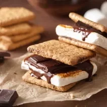 Fotografia de duas peças de s'mores sobre um papel manteiga. Ao fundo tem alguns pedaços de chocolate e alguns biscoitos empilhados, sobre uma mesa de madeira.