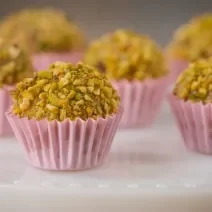 Fotografia de seis forminhas de cor rosa com brigadeiro de pistache enrolado no pistache triturado. Os brigadeiros estão em um apoio branco decorativo.