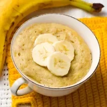 Fotografia em tons de amarelo com uma xícara de café ao centro. Dentro da xícara existe uma receita de bolo de banana. Ao lado existe uma banana inteira em cima de um pano amarelo