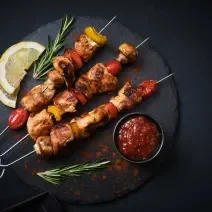 Fotografia vista de cima de três espetinhos, um do lado do outro, de carnes intercaladas com legumes. Os espetos estão sobre um apoio preto redondo, ao lado de duas fatias de limão siciliano, um molho vermelho em um recipiente menor preto e alecrim.