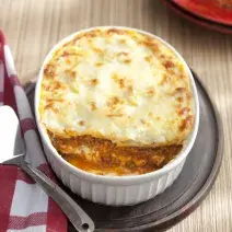 Fotografia em tons de branco e marrom, ao centro travessa branca com lasanha gratinada sobre tábua de cozinha, lado esquerdo guardanapo xadrez branco e vermelho, em bancada de madeira. Ao fundo, pedaço de lasanha em prato vermelho.