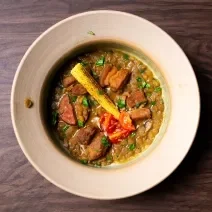 Fotografia em tons de marrom com um prato cinza ao centro. Em cima do prato existe uma receita de lentilha com carne de porco, tomates e mini milhos