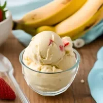 Foto da receita de Sorvete de Banana com Morango. Observa-se uma tacinha de vidro com as bolas de sorvete dentro, na cor amarela e com pedacinhos de morango. Foto gerada por IA.