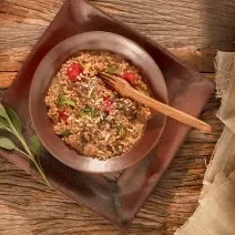 Foto em tons de marrom da receita de risoto ao funghi com aveia corte rústico servida em um prato fundo marrom sobre uma base retangular de mesma cor em cima de uma mesa de madeira