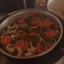 Fotografia de uma panela grande com moqueca com tomates, pimentões e cebolas em rodelas por cima. A panela está dentro de uma outra panela maior.