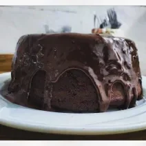 Fotografia em tons de preto em uma mesa de madeira, um pano vermelho, um prato branco raso redondo com o bolo de chocolate com cobertura de chocolate em cima.