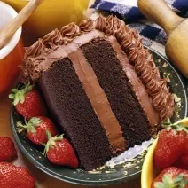 Fotografia de uma fatia de bolo com massa e recheio de chocolate, morangos por toda a parte em um prato desenhado de cor escura, apoiado em uma toalha de mesa azul e branca, quadriculada. Ao lado do bolo, dois potes e um rolo de massa.