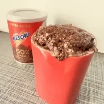 Fotografia de uma caneca vermelha com bolo de chocolate com grãos de aveia. Ao fundo tem uma embalagem de nescau 60% cacau.