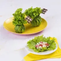 Fotografia em tons de branco, amarelo e verde de uma bancada branca vista de cima, contém um pano amarelo e um prato branco com a trouxinha de alface e ao lado um prato amarelo com duas trouxinhas de alface