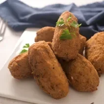 Fotografia de vários bolinhos de bacalhau fritos sobre um apoio branco, ao lado de um pano azul de tom escuro e um garfo.