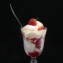 Fotografia de uma taça de vidro com merengue e morangos, de forma intercalada. E dentro do recipiente, uma colher de sobremesa apoiada, sobre um fundo preto.