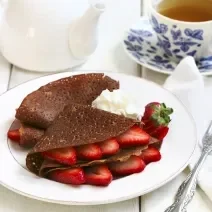 Fotografia de panquecas de chocolate com morango, e um pouco de chantilly ao lado, dentro de um prato branco raso. Ao redor , um garfo sobre uma toalha branca, um bule branco e uma xícara de chá em cima do prato pequeno, ambos são azuis e brancos.