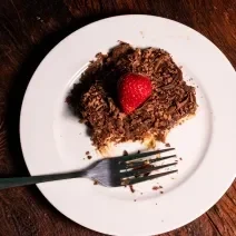 Fotografia vista de cima de um pedaço de pavê com mousse e chocotrio. Por cima tem ganache, raspas de chocolate e morango. O pavê está sobre um prato branco de vidro com um garfo.