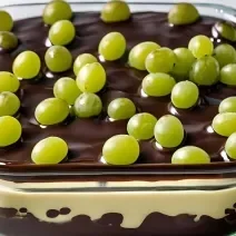 A fotografia mostra um refratário com camadas de uvas verdes sem sementes, cobertas por um creme branco espesso e finalizadas com uma ganache de chocolate ao leite brilhante.