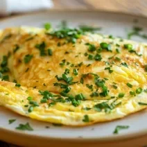 Fotografia de uma omelete rápida sobre um prato de vidro raso, e por cima da omelete tem cebolinha picada salpicada. O prato está sobre uma mesa de madeira.