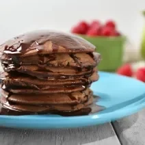 Fotografia de algumas panquecas de chocolate empilhadas em um prato azul claro, e sobre as panquecas tem uma calda de chocolate escorrendo. Ao fundo em algumas framboesas dentro de um recipiente verde, e espalhadas sobre a mesa de madeira branca.