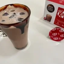 Fotografia em tons de vermelho com uma caneca de vidro ao centro. Dentro da caneca existe uma receita de Iced Mocha feita com Dolce Gusto Kitkat. Nas bordas da caneca existe uma calda de chocolate