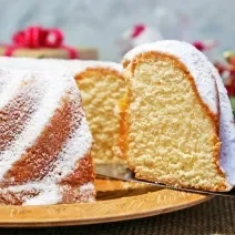 Fotografia de um bolo de massa branca de leite com um pedaço em destaque e cobertura de açúcar de confeiteiro, sobre um apoio reto de tom claro.