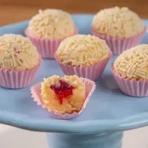 Fotografia de seis forminhas rosa com brigadeiro branco, passado no granulado branco, com recheio de frutas vermelhas. Os brigadeiros estão apoiados em um apoio azul. O brigadeiro da frente está mordido, mostrando a massa e o recheio.