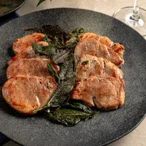 Foto de um prato preto visto de cima, com a receita de mignon suíno com manteiga e sálvia, em fatias, sobre uma mesa em que se vê a parte de baixo de uma taça de vinho e alguns talheres