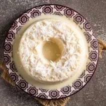 Fotografia vista de cima de um prato branco com desenhos de tom escuro com um pudim de coco com raspas de coco por cima. O prato está apoiado sobre um pano marrom com rebarbas decorativas.