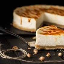 Fotografia de uma fatia de cheesecake com caramelo salgado por cima de uma espátula de prata, sobre uma mesa preta. No fundo tem o resto da cheesecake sobre um apoio preto.