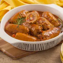 Fotografia em tons de amarelo em uma bancada de madeira clara, um paninho amarelo, uma tábua de madeira, com um recipiente branco redondo com vários bifes à role dentro super suculento.