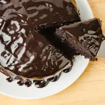 Fotografia em tons de marrom e branco de uma bancada marrom vista de cima, contém um prato redondo e branco com um bolo de chocolate com cobertura de chocolate e uma fatia sendo retirada por uma espátula
