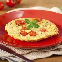 Fotografia em tons de vermelho em uma bancada de madeira, um pano bege, um prato vermelho redondo com a omelete com tomates-cereja em cima dele. Ao fundo, tomatinhos para decorar e ao lado, garfo e faca.