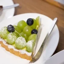 Fotografia de uma fatia de torta recheada com leite MOÇA e uvas verdes por cima, com três uvas roxas. Por cima da torta tem açúcar de confeiteiro polvilhado, e ao lado, um garfo sobre um prato branco, que está sobre uma mesa de madeira.