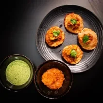 Fotografia vista de cima de um prato preto raso com quatro unidades de pani puri com grão de bico e coentro. Ao lado tem dois molhos em recipientes diferentes, um molho de iogurte e o outro, um molho de manga. Tudo está sobre uma mesa preta.