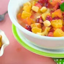 Fotografia em tons de rosa, branco e verde de uma bancada rosa vista de cima, um pano verde com bolinhas brancas e acima um prato branco e verde e por cima do prato um recipiente branco e redondo com frutas. Ao lado um pote branco com frutas.