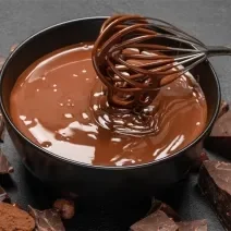 Imagem de um pequeno recipiente contendo uma calda de chocolate cremosa e derretida, em tons de marrom. Um batedor de arame mexe mistura. Ao fundo, barras de chocolate.