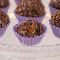 Fotografia em tons de roxo com várias forminhas de brigadeiro roxas ao centro. Dentro de cada forminha existe um brigadeiro de chocolate meio amargo recheado com um creme de caramelo salgado.