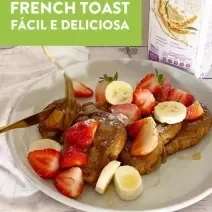 Foto em tons claros da foto da receita de french toast servida com bananas, morangos e uma calda sobre um prato de porcelana branco. Ao fundo, há uma embalagem de bebida vegetal de aveia natures heart