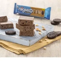 Fotografia mostra quadradinhos de paçoca em tom de chocolate, ao lado amendoins e biscoitos. Ao fundo um pacote de biscoito de amendoim.