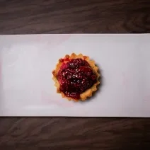 Fotografia em tons de branco com um prato branco ao centro. Em cima do prato existe uma receita de Tartallete coberto com uma geleia de frutas vermelhas e pedaços de frutas vermelhas.