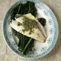 Fotografia em tons de branco em uma bancada de madeira de cor branca. Ao centro, um prato contendo o peixe.