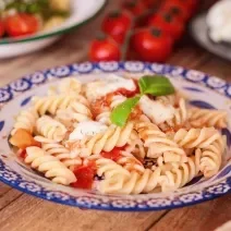 Fotografia de macarrão parafuso com tomate fresco e pinhole, queijo ralado e duas folhas de manjericão por cima. O macarrão está em um prato branco e azul, o qual está sobre uma mesa de madeira.