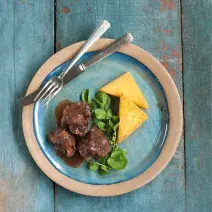 Fotografia em tons de azul em uma bancada de madeira azul com um prato redondo raso rosa claro com azul e a rabada, a polenta e salada de agrião em cima dele.