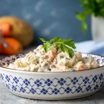 Fotografia de uma salada de maionese que está em um recipiente fundo com desenhos azuis. Tem um ramo de salsinha apoiado na salada. O recipiente está apoiado em uma bancada de textura parecida com gesso, ao fundo tem frutas.