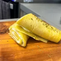 Fotografia em tons de amarelo com uma tábua de madeira ao centro. Em cima da tábua existe uma crepioca recheada com creme de ricota e Ninho Adulto