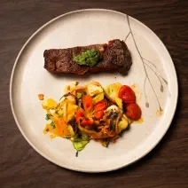 Fotografia vista de cima de um bife ancho com pesto de cerefólio e salada de cogumelos ao lado. A receita está em um prato grande, branco e raso, o qual está sobre uma mesa de madeira.