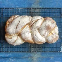 Fotografia vista de cima de um pão doce trançado de leite MOÇA e recheio de beijinho. Por baixo do pão tem uma grade preta que está sobre uma mesa de madeira azul.